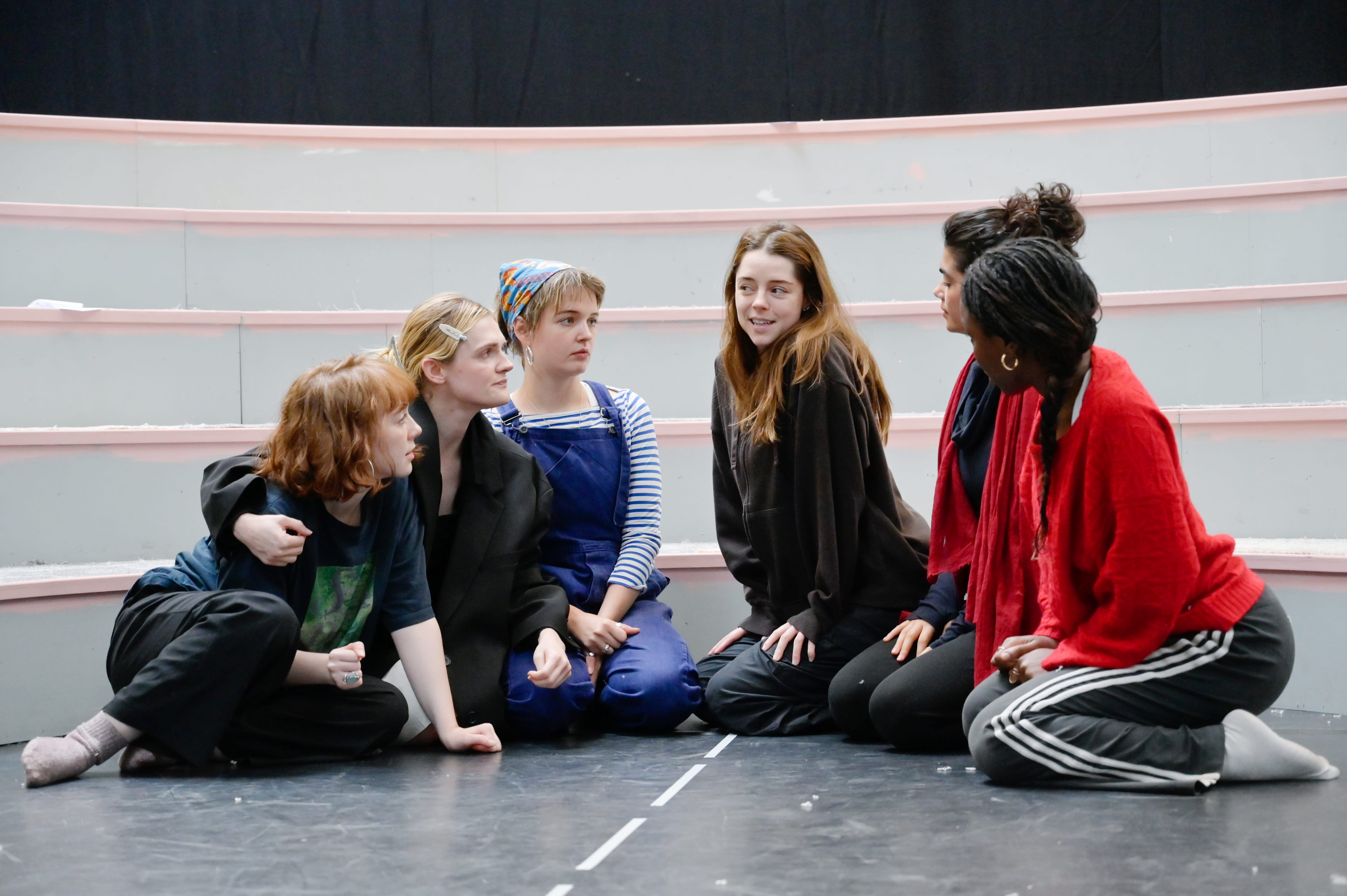 A group of women are sitting on the floor in a semi-circle. They are all looking at one girl in the middle who is speaking.