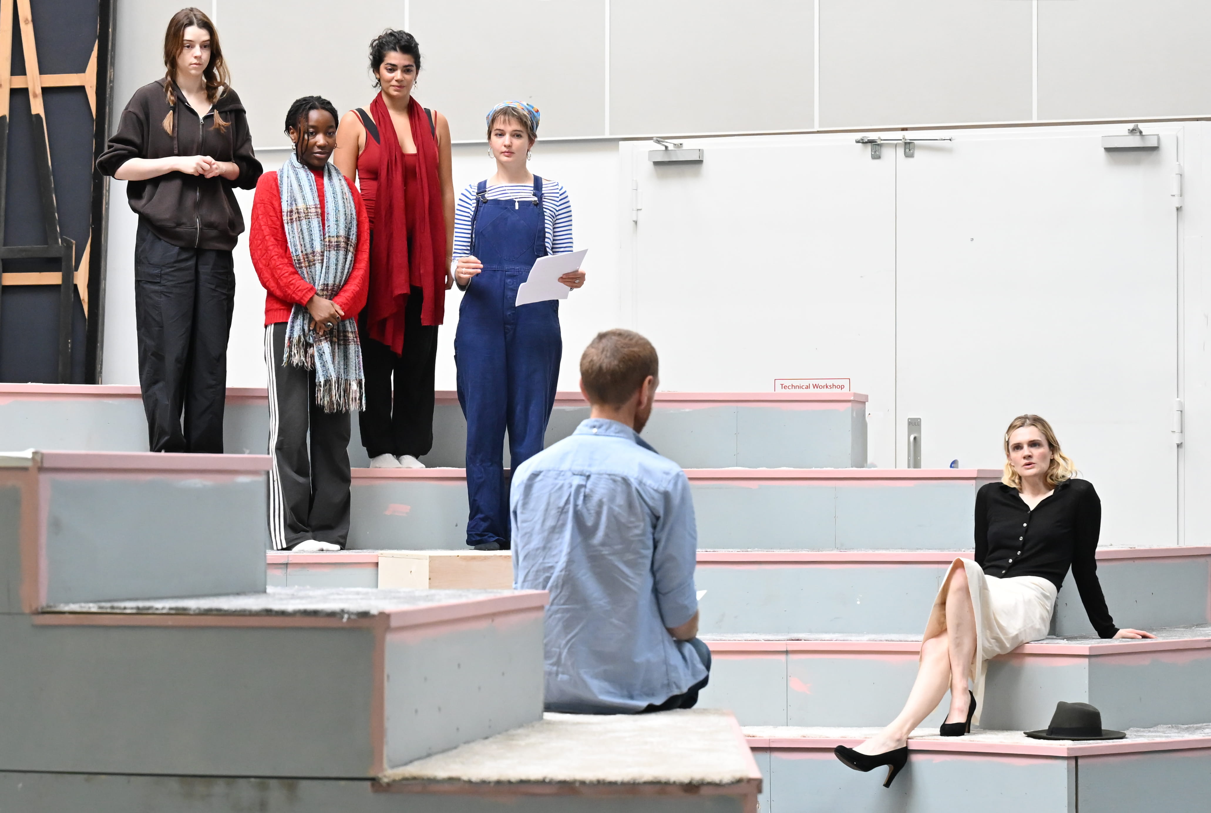 Four women stand on a set of steps, in an amphitheatre layout. At the bottom of the steps, a man and woman sit facing each other. The man has his backs to the camera.