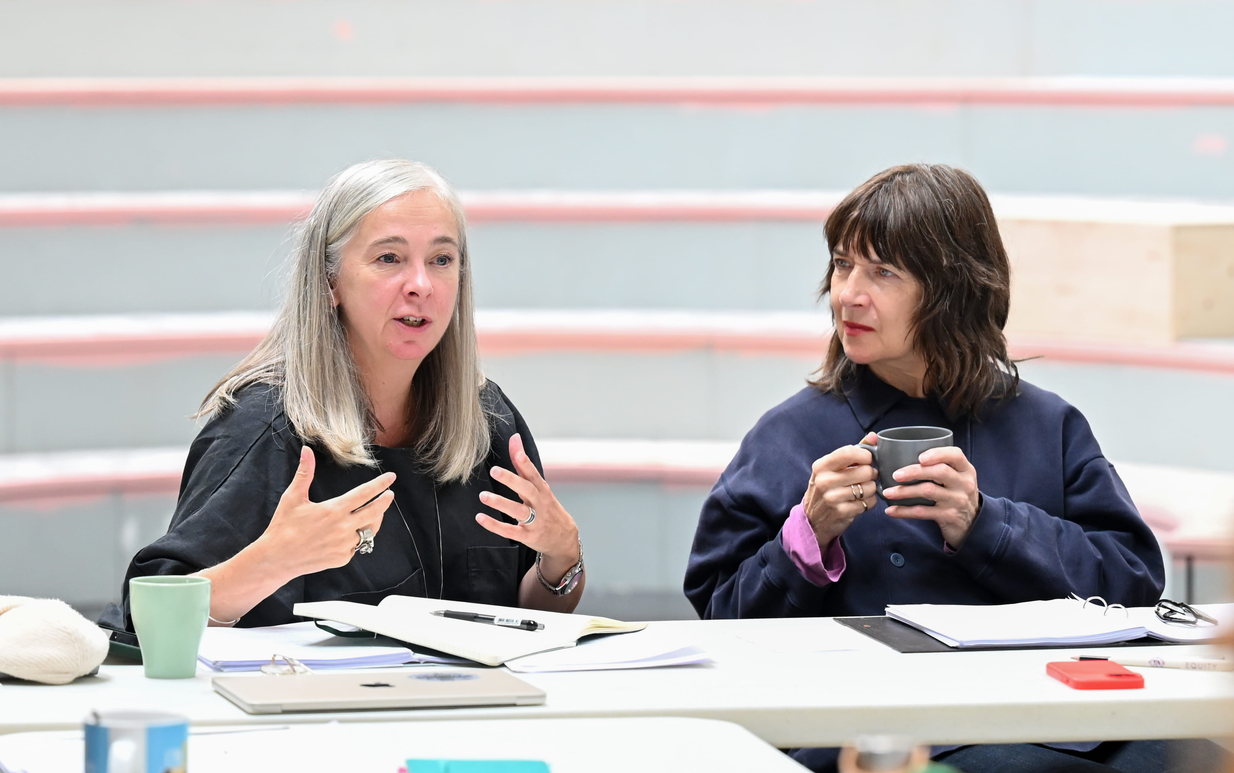 Two women are sitting next to each other at a table. One is speaking, and the other is looking at her whilst holding a cup of coffee.